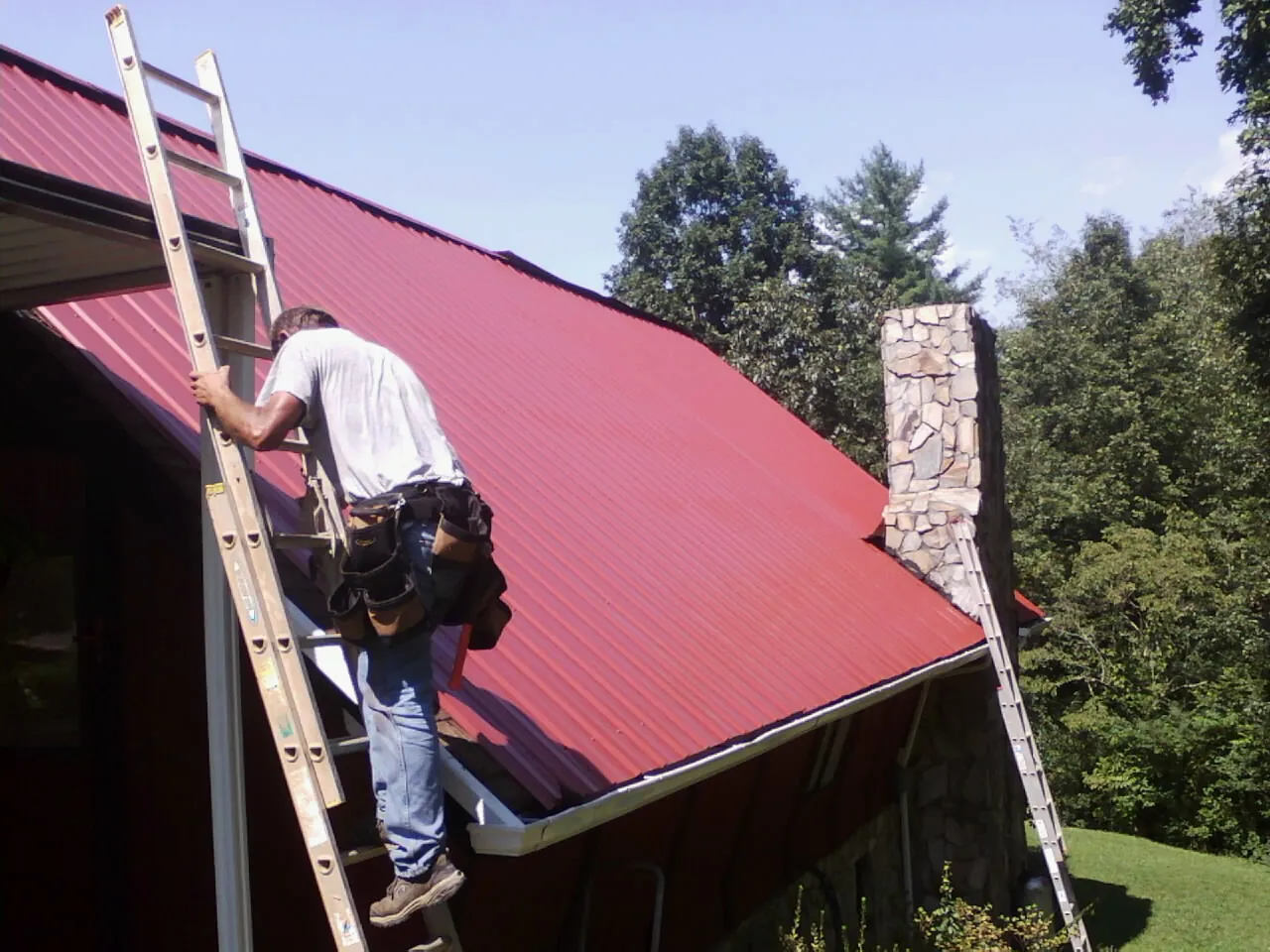 Roofer on a ladder beside a red metal roof and stone chimney — Soffit & Fascia Repair by Roofing Brothers in Volcano